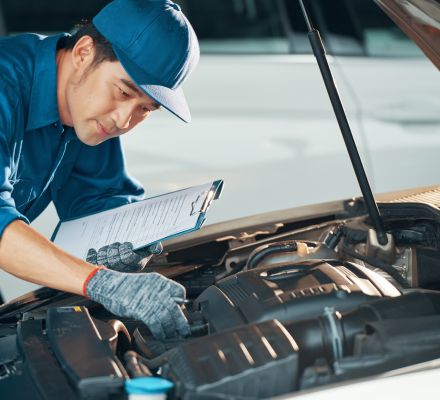 Mechanic inspecting a car battery while holding a clipboard.